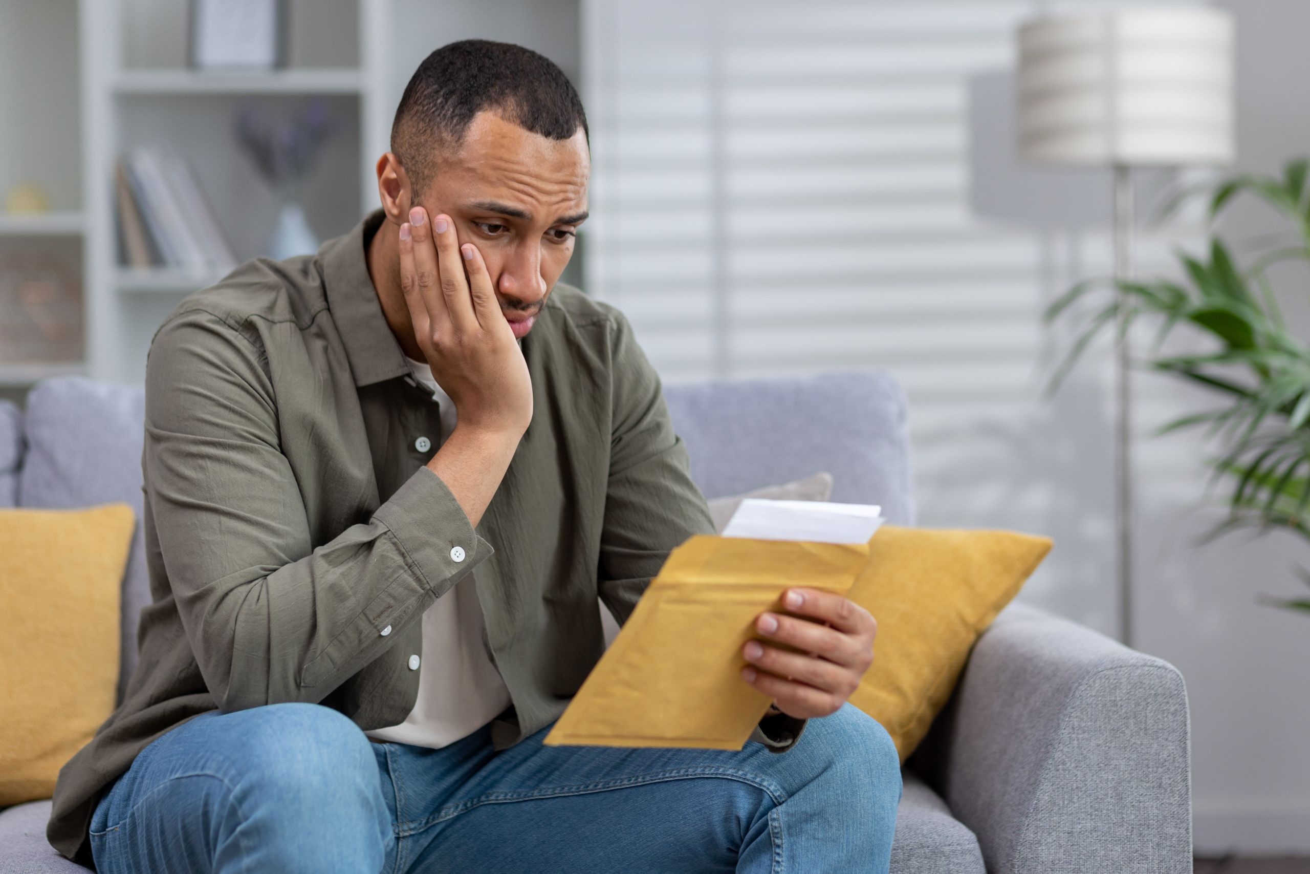 Worried man sitting on couch reading a letter from an envelope, representing IRS notice, tax debt, or potential bank levy concern.