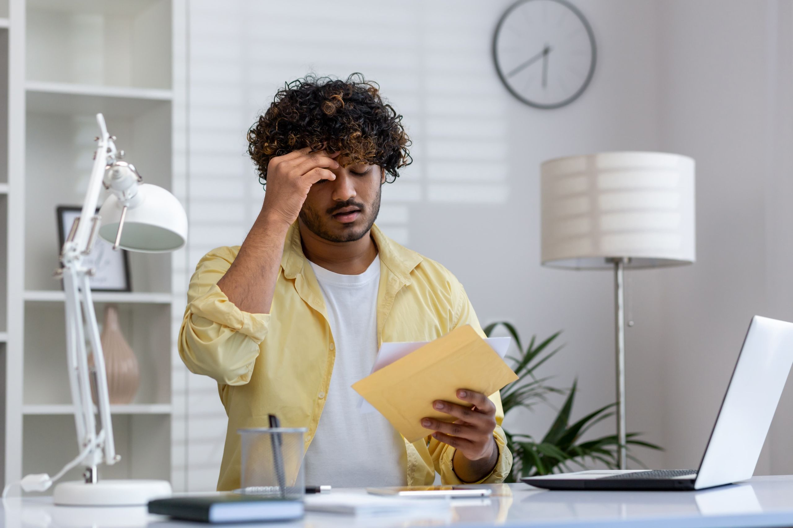 Stressed man sitting at his home desk holding an envelope and reading an IRS notice, representing the anxiety of owing the IRS more than $10,000